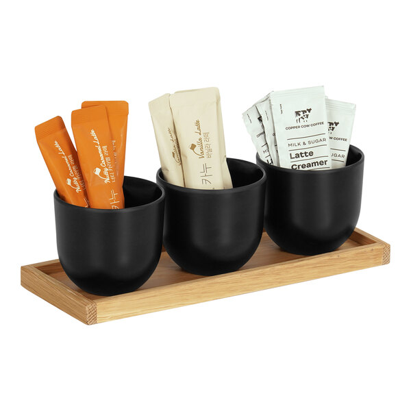 A Cal-Mil oak condiment organizer with three black round melamine jars filled with coffee and tea packets on a wooden tray.