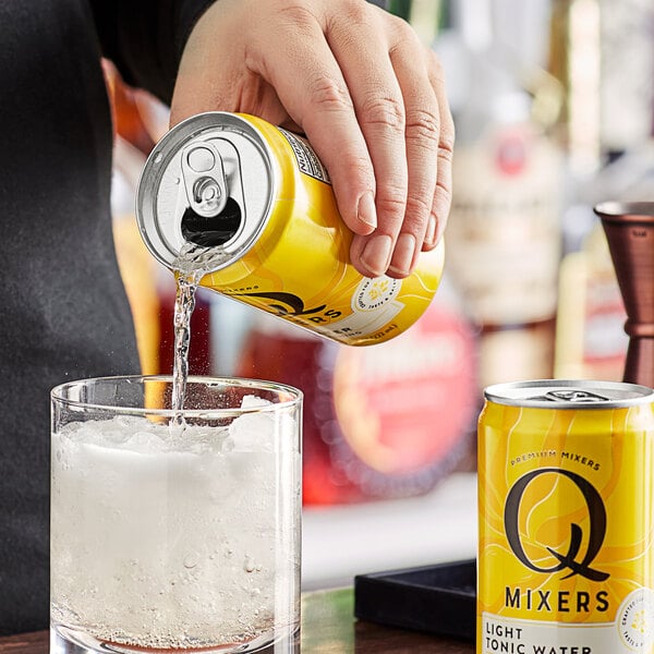 A hand pouring Q Mixers Light Tonic Water from a yellow can into a glass with ice.