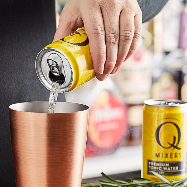 A hand pouring Q Mixers Tonic Water from a yellow can into a copper cup.