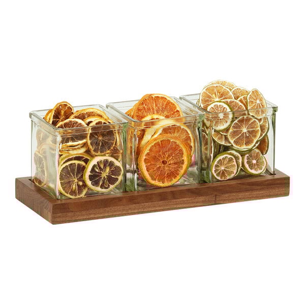 A Cal-Mil walnut condiment organizer with three square glass jars filled with citrus slices.