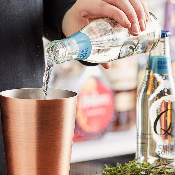 A hand pouring Q Mixers Premium Club Soda from a clear bottle into a copper cup.