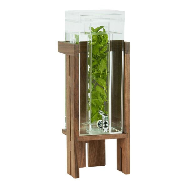 A Cal-Mil glass beverage dispenser with a plant inside on a wooden stand.