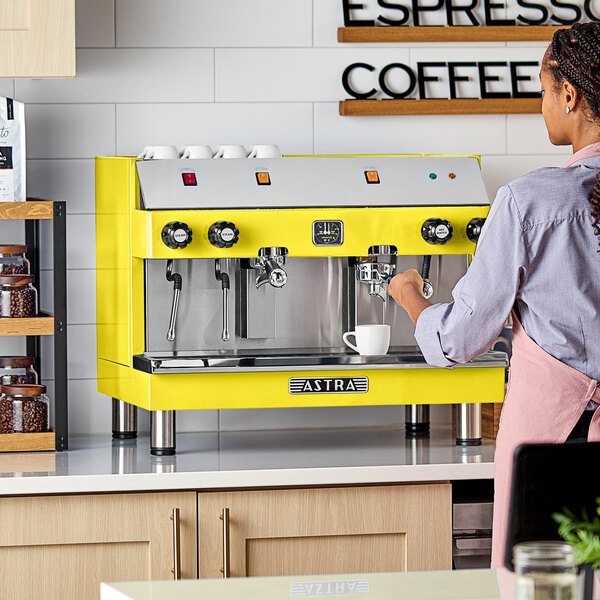 A yellow Astra Mega II semi-automatic espresso machine on a countertop with a person preparing coffee.
