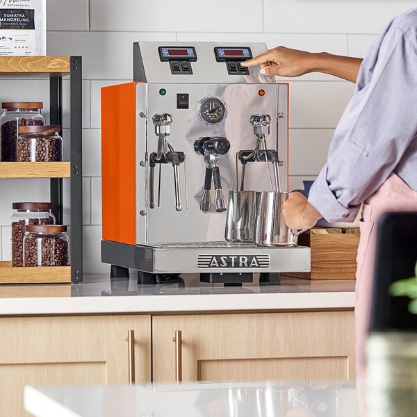 An orange Astra automatic milk and beverage steamer with two wands on a countertop.