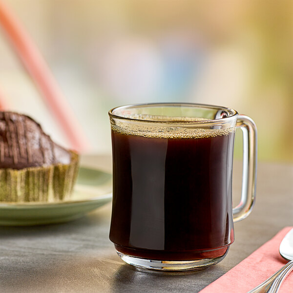 A glass mug of Folgers Colombian ground coffee on a table with a blurry cupcake.
