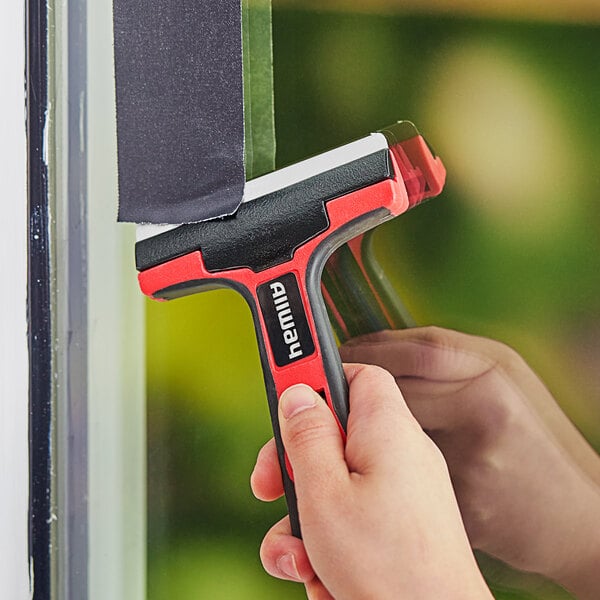 A person using an Allway Tools glass and tile scraper with a red and black handle to clean a window.