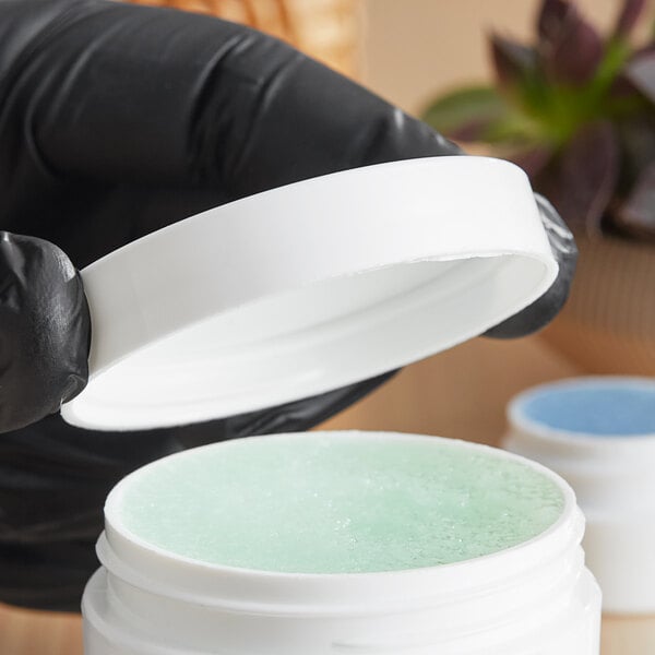 A hand in a black glove holding a white jar with a 58/400 smooth white lid filled with green gel.