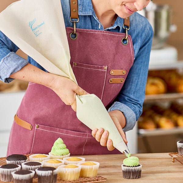 A woman in a red apron using an Ateco plastic-coated canvas pastry bag to pipe green frosting onto a cupcake.
