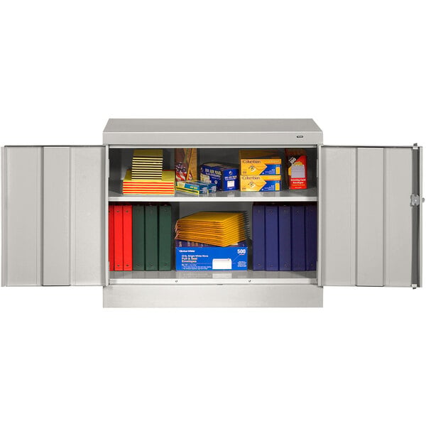 A Tennsco light gray metal storage cabinet with solid doors open to reveal shelves with different colored boxes and folders.