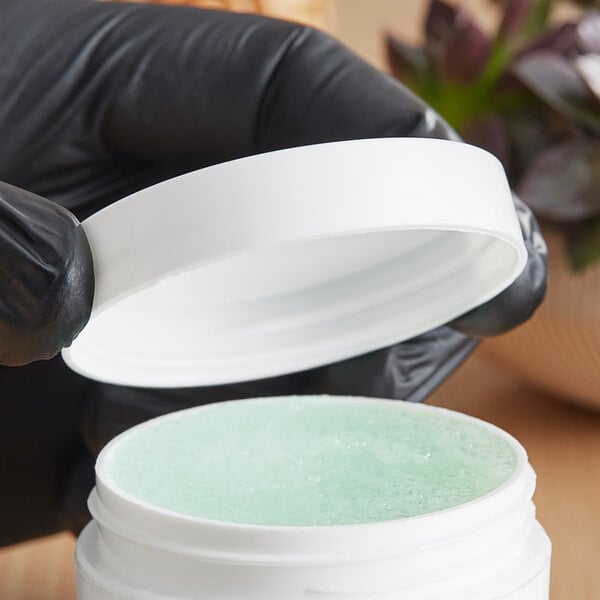 A hand in a black glove holding a white jar with a green substance and a white 58/400 smooth unlined lid.