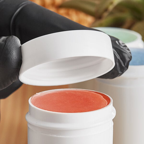 A hand with black gloves holding a white container with red liquid and a white lid.