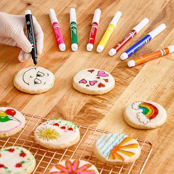 A hand using an Ateco food coloring marker to draw a rainbow on a cookie.