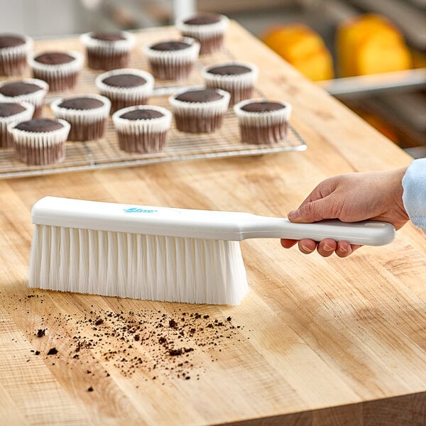 A white Ateco counter duster brushing crumbs off a wood table with cupcakes in white wrappers.