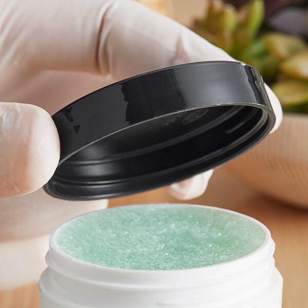 A hand holding a white jar with a black smooth lid filled with blue gel.