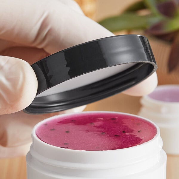 A hand holding a white container of pink liquid with a black lid.