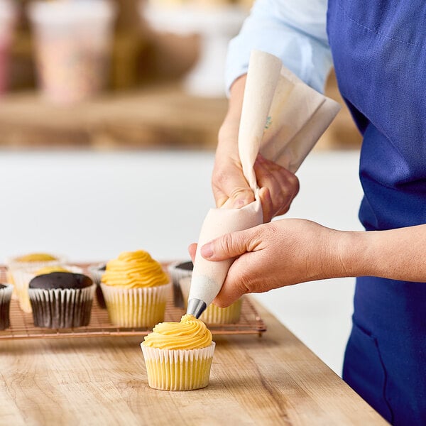 A person decorating a cupcake with a yellow swirl using an Ateco plastic-coated canvas pastry bag.