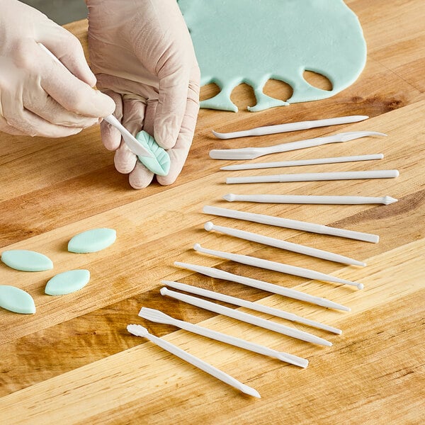 A gloved hand uses white plastic sticks from the Ateco Mini Sculpturing Tool Set to cut blue foam.