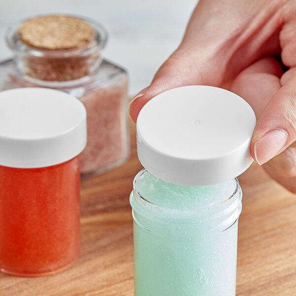 A hand holding a 38/400 smooth white lid over a small glass jar.