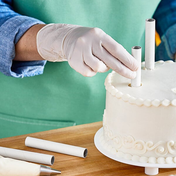 A hand in a white glove placing an Ateco parchment-coated paperboard dowel on a white cake.