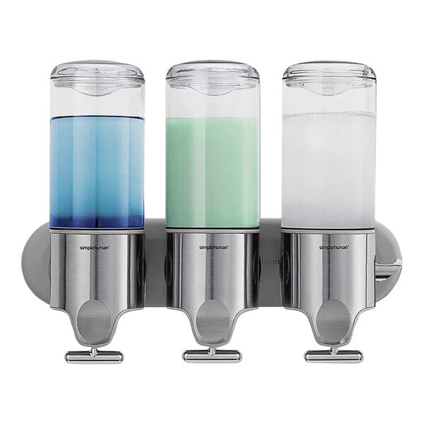 A simplehuman stainless steel triple soap dispenser on a wall with three glass bottles with blue, white, and green liquid soap.