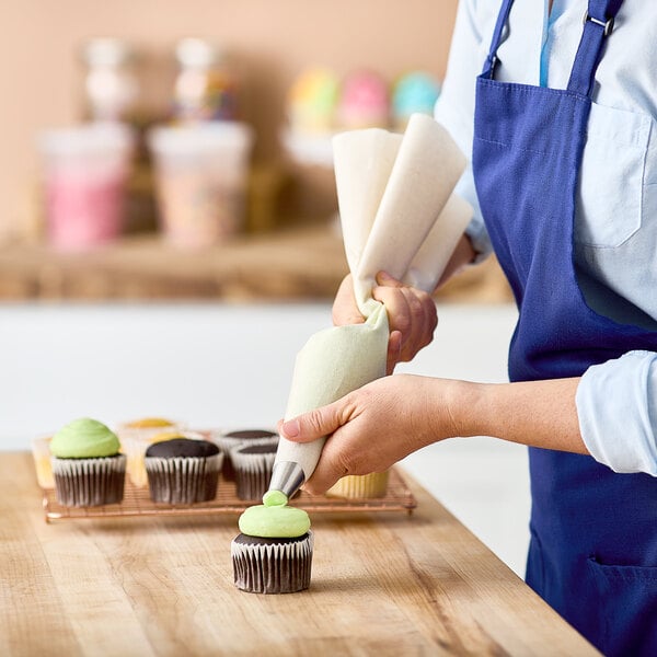 A person in a blue apron using an Ateco plastic-coated canvas pastry bag to decorate a cupcake.