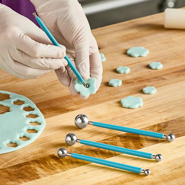 A close-up of a gloved hand using an Ateco blue two-sided modeling ball tool.