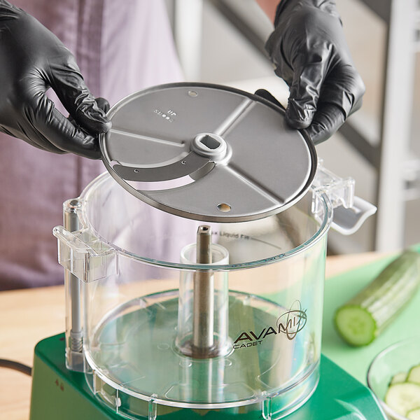 A gloved hand uses an AvaMix 5/32" slicing disc in a food processor to slice a cucumber.