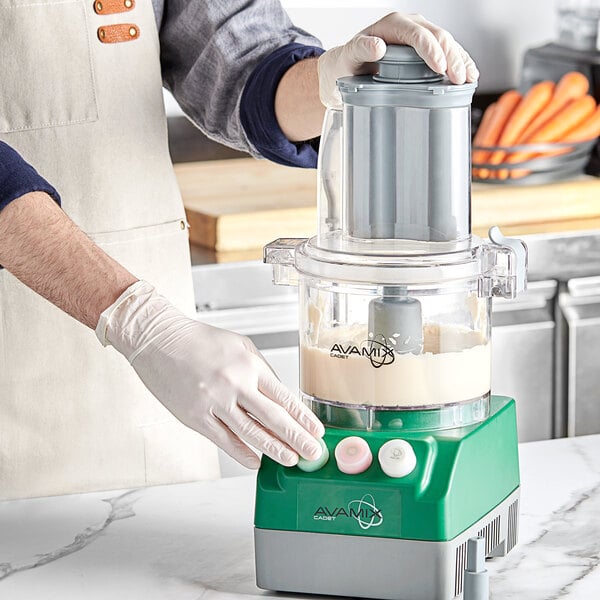 A person in a white apron and gloves using an AvaMix Cadet food processor to chop carrots.