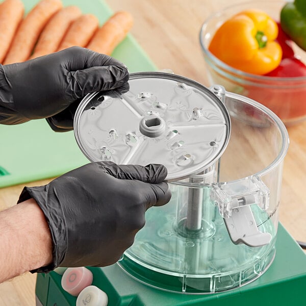 A person wearing black gloves uses an AvaMix grating disc to shred carrots in a food processor.