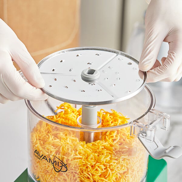 A gloved hand uses an AvaMix Cadet grating / shredding disc in a food processor.