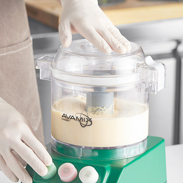 A gloved hand using an AvaMix clear plastic batch bowl with flat lid and "S" blade to mix white liquid.