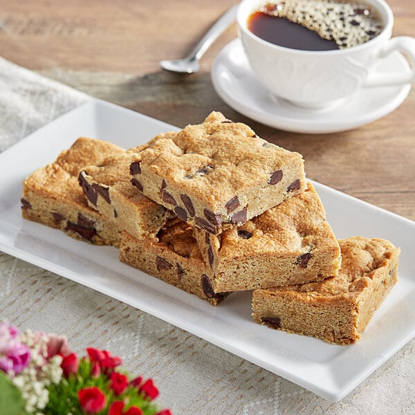 A Root Nine Baking Co. vegan blondie tray on a table with a plate of brownies and a cup of coffee.