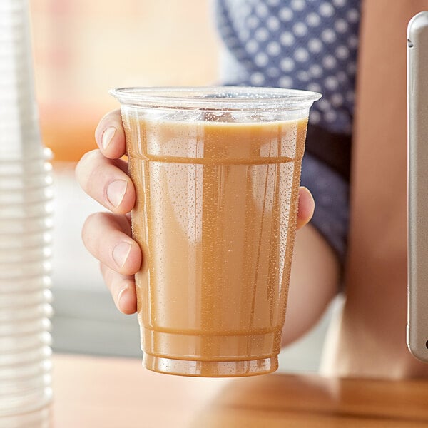 A hand holding a Choice clear plastic cup of brown liquid.