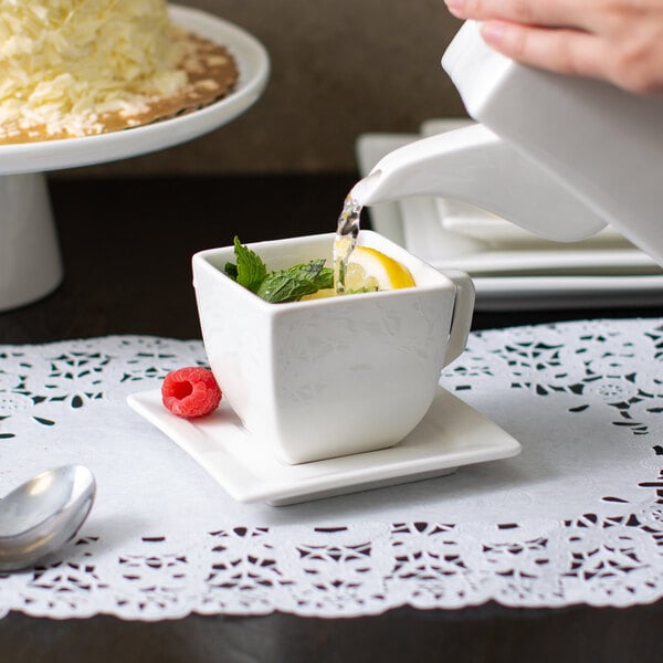 A person pouring water from a faucet into a white square porcelain cup of tea with a lemon and mint leaves.