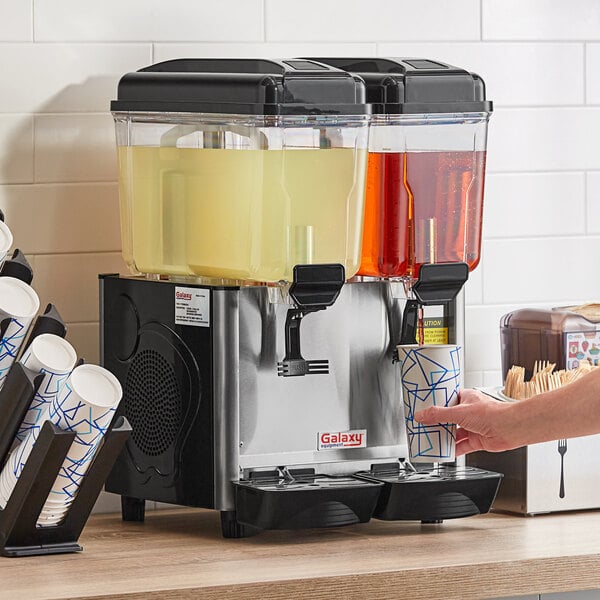 A hand holding a cup of yellow beverage next to a Galaxy refrigerated beverage dispenser.