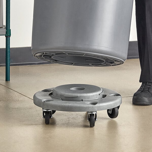 A person using a Lavex round grey dolly to move a large grey trash can.