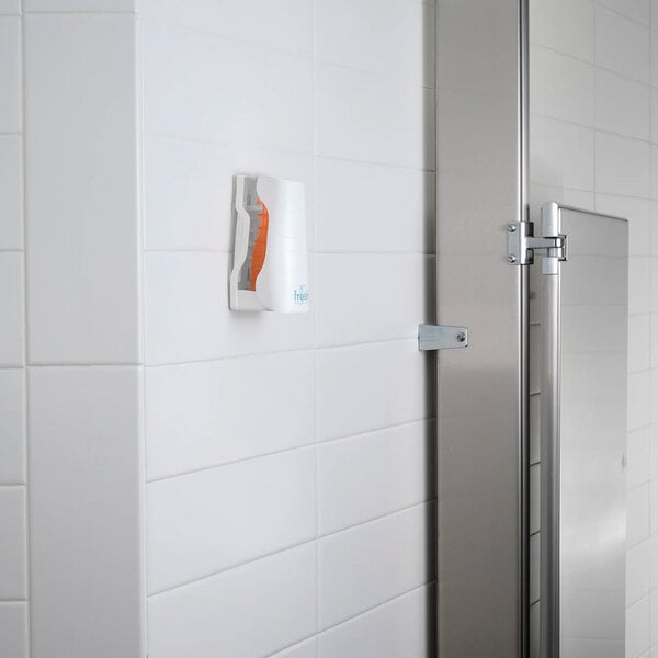A wall-mounted passive air freshener refill with a mango scent in a white holder on a tiled bathroom wall.