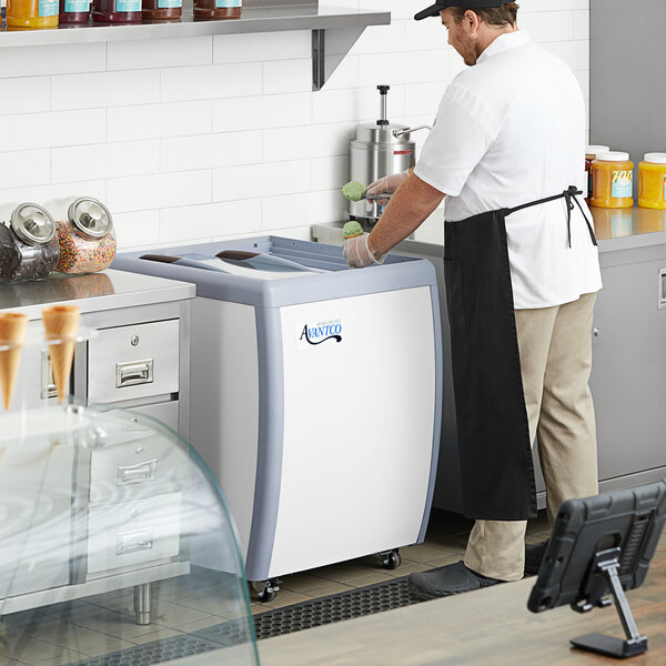 An Avantco ice cream dipping cabinet on a counter with a glass dome over it, being used by a man in a white shirt and black apron.