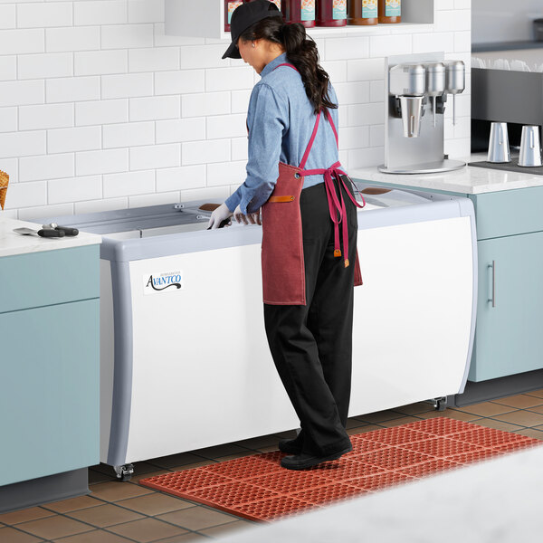 A woman wearing a blue apron and gloves scooping gelato from an Avantco gelato dipping cabinet.