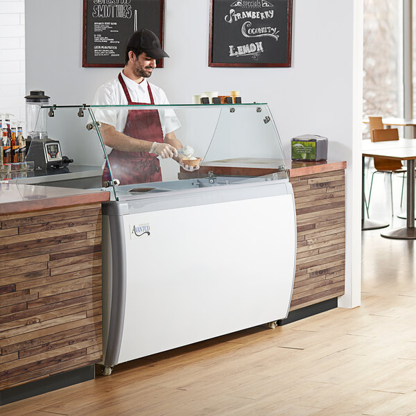 An Avantco ice cream dipping cabinet with a clear top on a counter with a man in a white hat and apron.