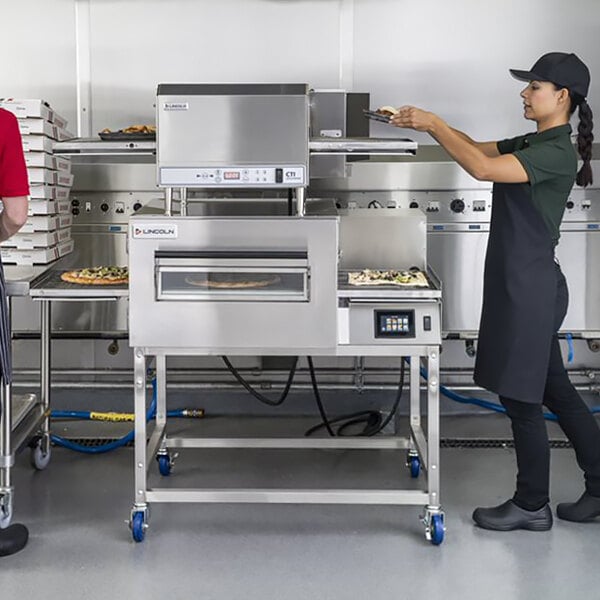 A woman and a man using a Lincoln Impinger II conveyor oven to bake a pizza.