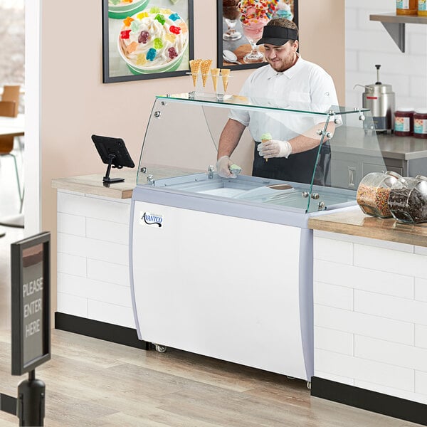 A man in a white shirt and black cap working at an Avantco ice cream dipping cabinet.