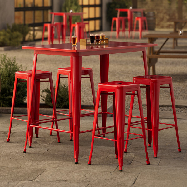 A Lancaster Table & Seating ruby red metal table with four backless barstools on an outdoor patio.