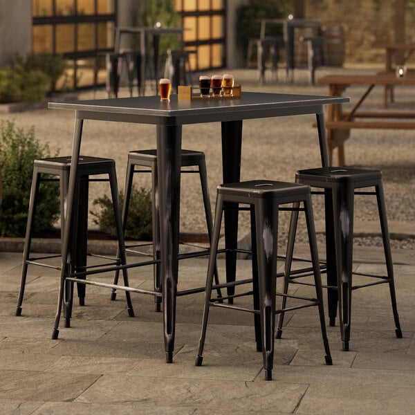 A Lancaster Table & Seating Alloy Series distressed onyx black bar table with four barstools outside.