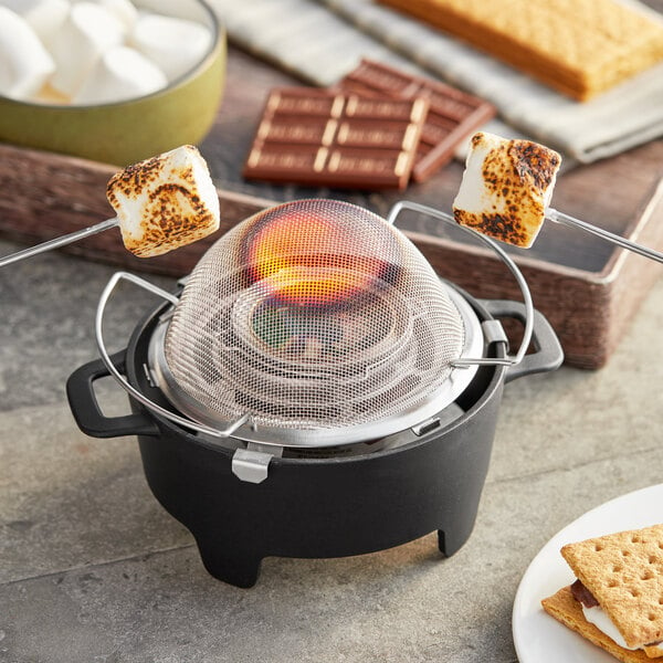 A small round black Sterno S'mores Maker Jr. with a mesh cover over a fire with marshmallows on it.