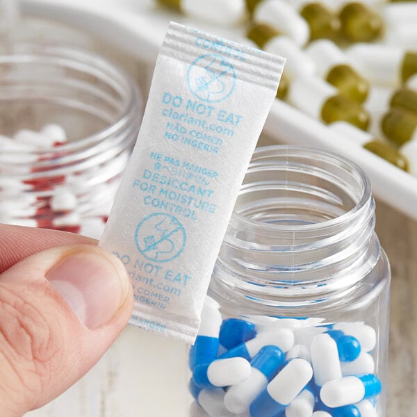 A hand holding a Clariant Sorb-It silica packet next to a jar of pills.