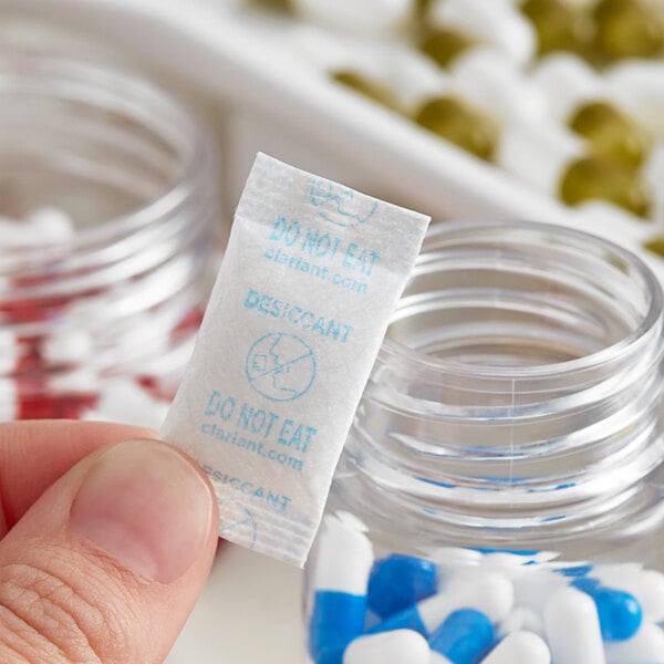 A hand holding a white Clariant Sorb-It silica packet over a jar of pills.