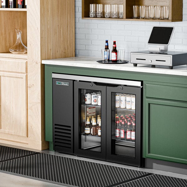 A True black glass door back bar refrigerator on a counter with bottles of beer inside.