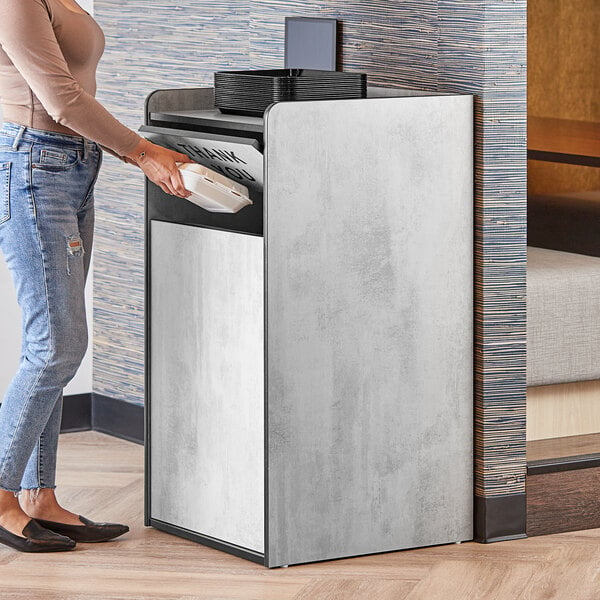 A woman standing next to a white rectangular Lancaster Table & Seating waste receptacle with a "THANK YOU" swing door.