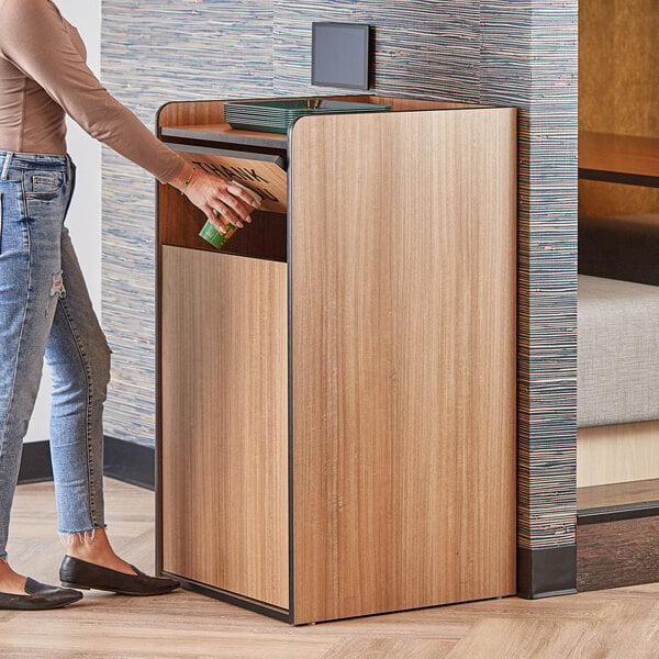 A woman putting a bottle of water into a Lancaster Table & Seating weathered walnut trash can with a "THANK YOU" swing door.